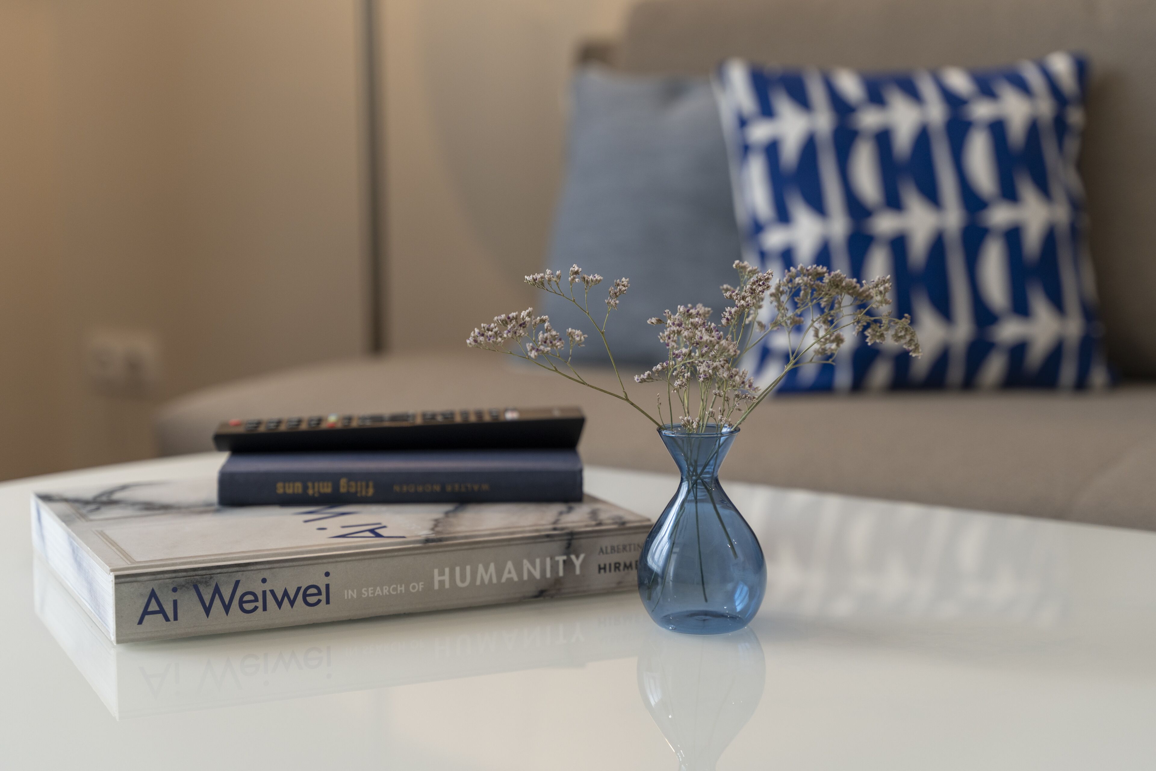 A white table with a blue vase containing dried flowers, a book titled 'Ai Weiwei: In Search of Humanity', and a remote control.