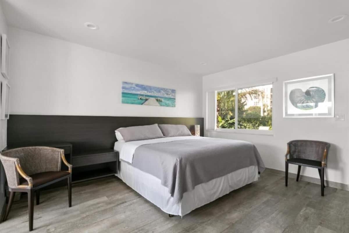 A modern bedroom with a bed, two chairs, a dark headboard, and artwork on the walls.
