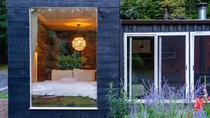 A modern cabin with a large glass window revealing a bed and a hanging light fixture. - Container Cabin in the Catskills [Field] (Saugerties)