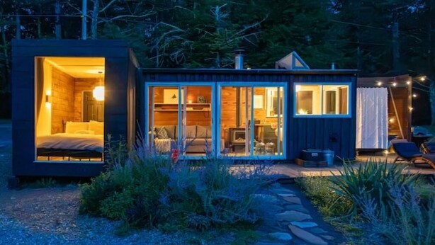 A modern cabin with a glass-enclosed living area, a visible bedroom, and a small outdoor seating area. - Container Cabin in the Catskills [Field] (Saugerties)