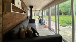 A cozy cabin interior with a wooden wall, a sofa with cushions, a fireplace, and a glass door leading to a deck with a view of greenery. - Container Cabin in the Catskills [Field] (Saugerties)