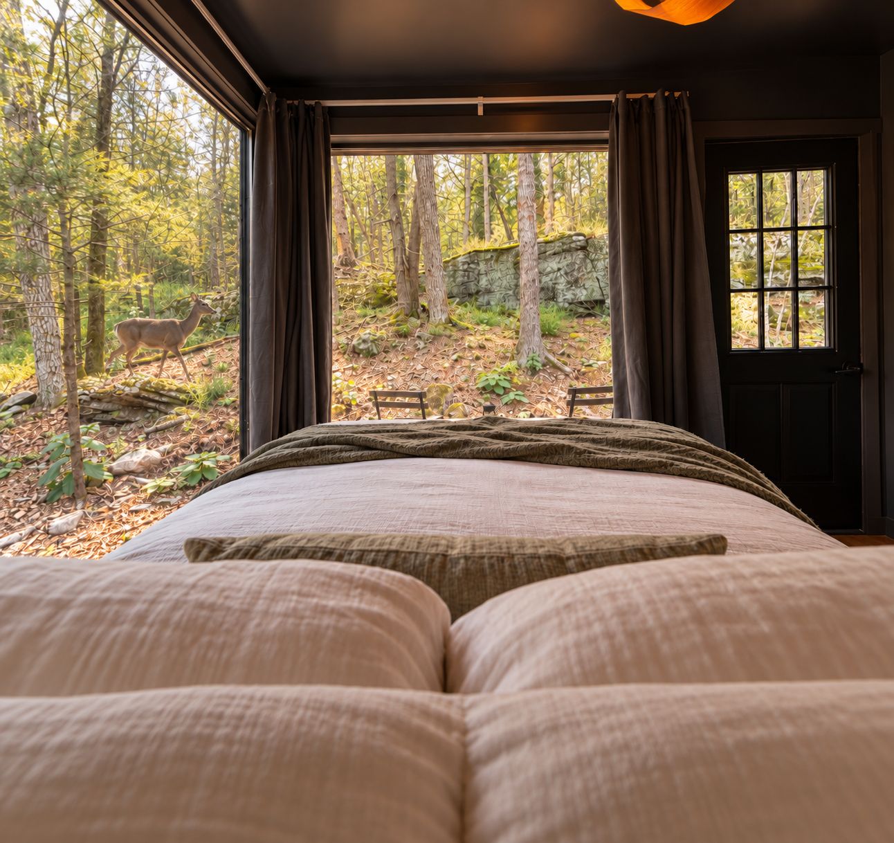 Une chambre avec un grand lit, une fenêtre avec des rideaux et une vue sur une forêt où l’on peut apercevoir un cerf.