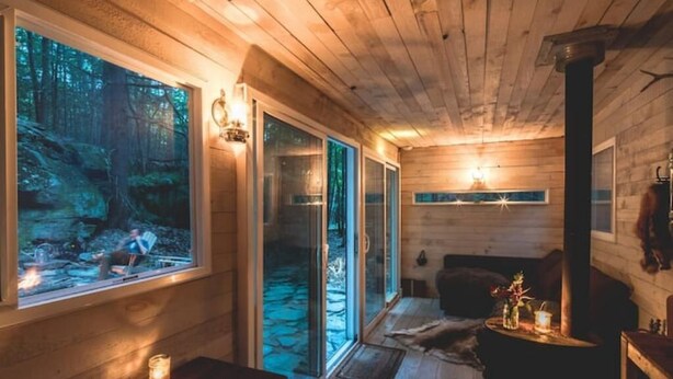 A cozy cabin interior with wooden walls, a fireplace, and a large window overlooking a forest.
