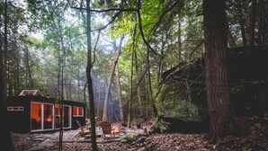 A cabin in a forest with a stone patio, a fire, and a chair.