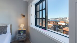 A bedroom with a large window, a bedside table with a lamp, and a view of buildings. - Myriverplace N.6 - PORTO Riverfront Apartment With Breathtaking Views (Vila Nova de Gaia)