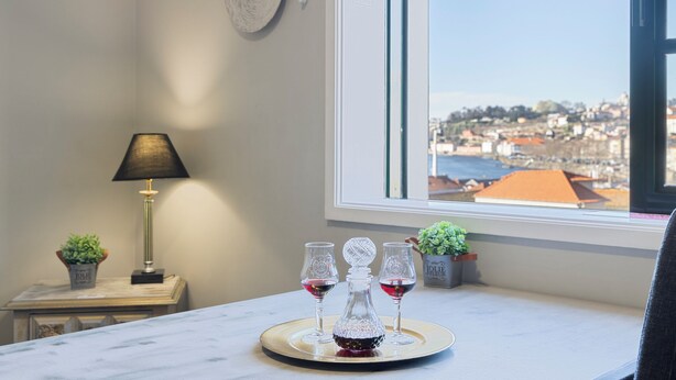 A room with a view of a cityscape through a large window, a marble table with two wine glasses and a decanter, a lamp, and a small plant on a side table. - Myriverplace N.6 - PORTO Riverfront Apartment With Breathtaking Views (Vila Nova de Gaia)