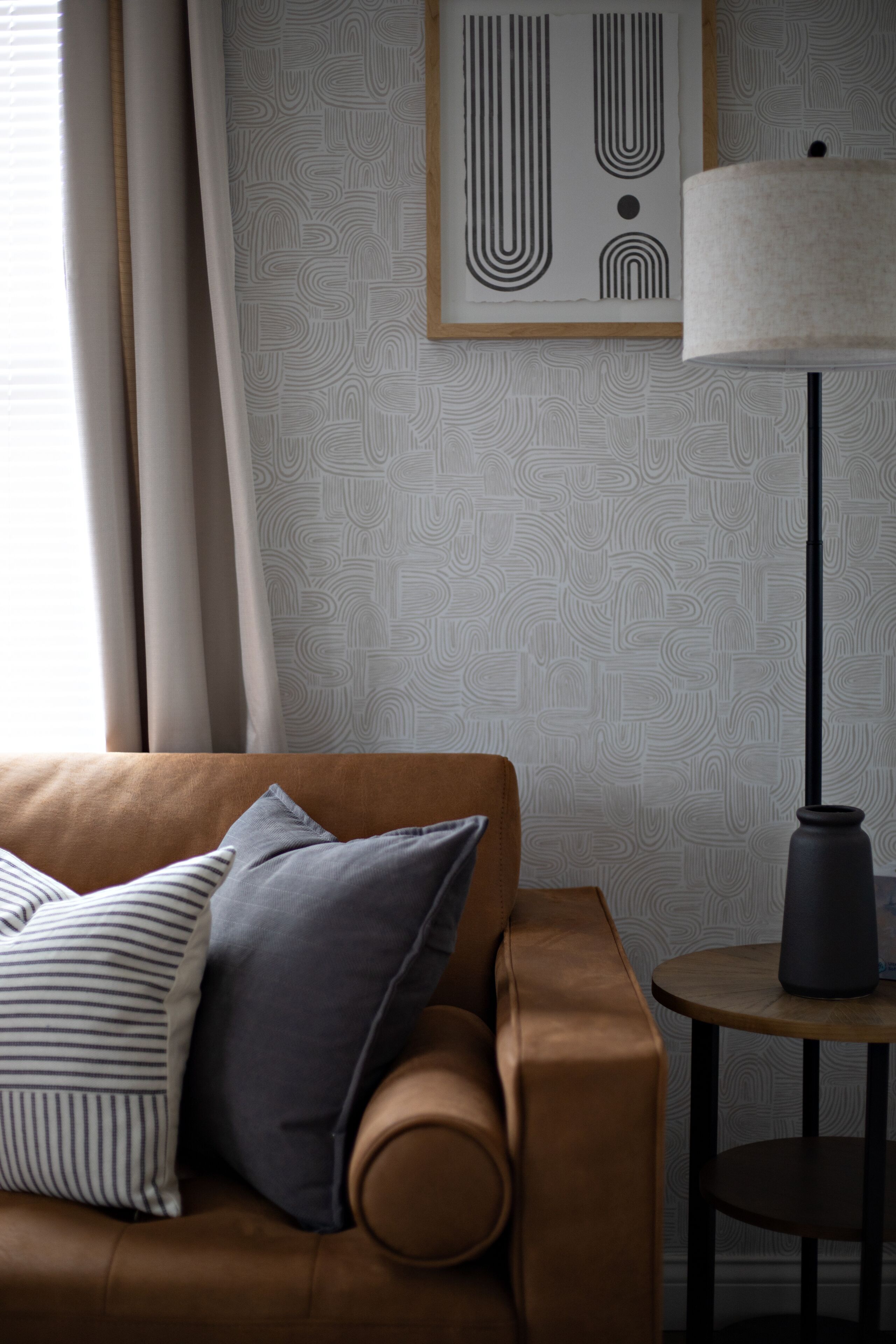 Een bruine leren bank met gestreepte en effen kussens, een ronde tafel met een lamp en behang met abstracte patronen.