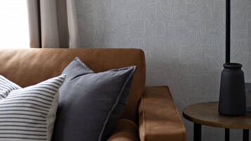 Een bruine leren bank met gestreepte en effen kussens, een ronde tafel met een lamp en behang met abstracte patronen.