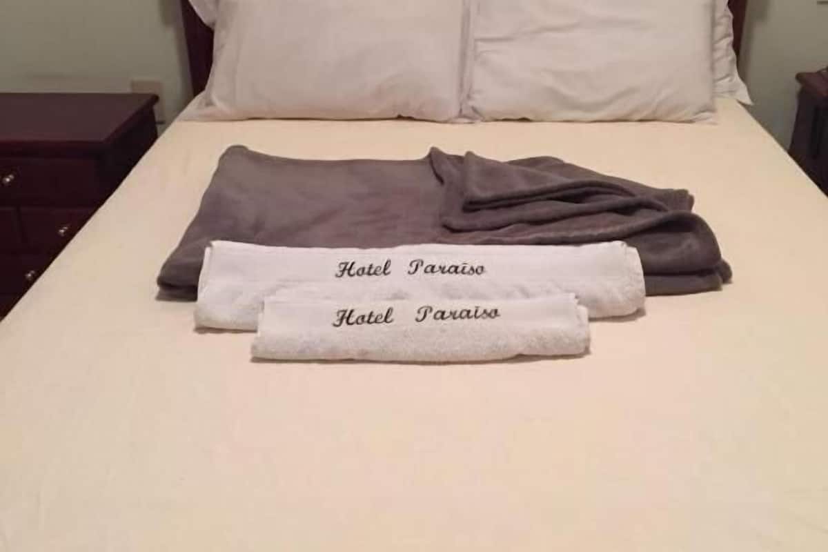 Un lit bien fait, avec un drap blanc, deux oreillers et une serviette pliée sur laquelle on peut lire le texte « Hôtel Paraiso ».