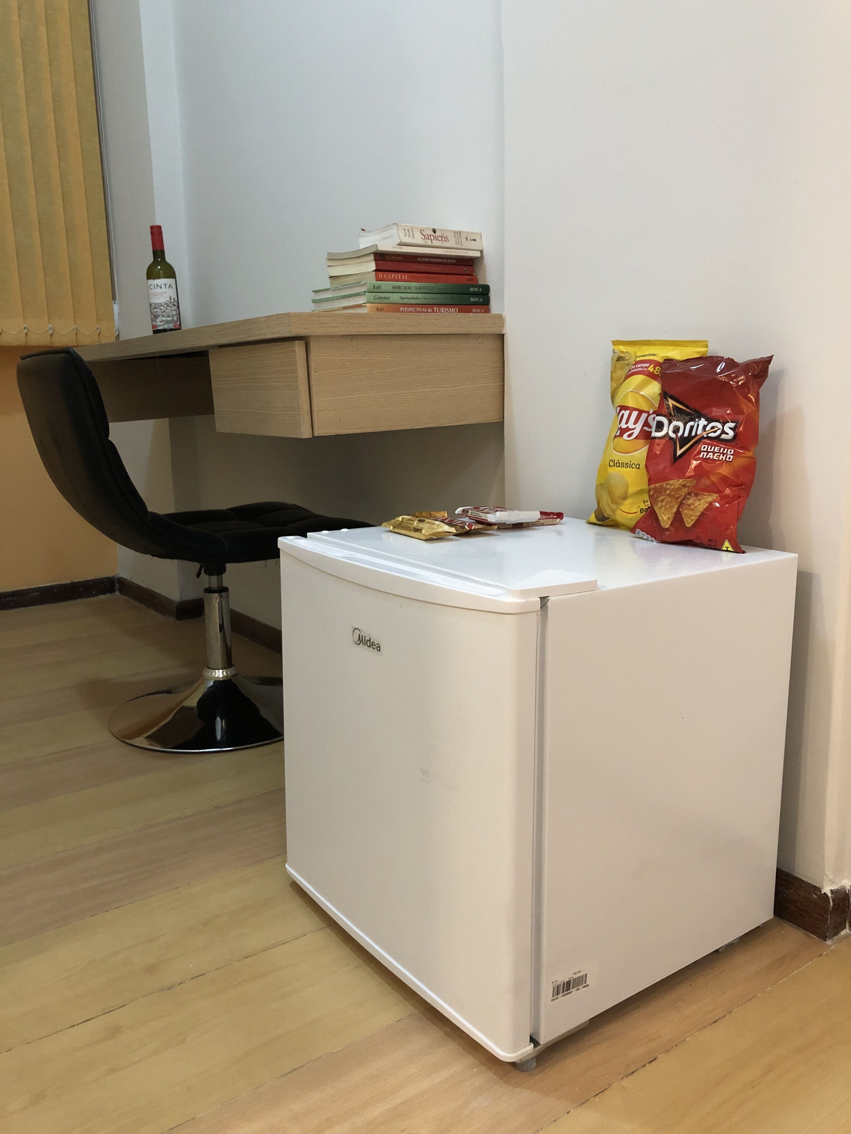 Un refrigerador blanco compacto con un congelador pequeño, un escritorio de madera con una silla, y una bolsa de papas.