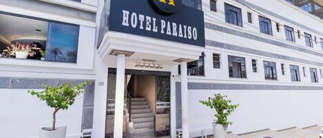 A hotel entrance with a sign reading "HOTEL PARAISO," a staircase leading inside, and two potted plants on either side.