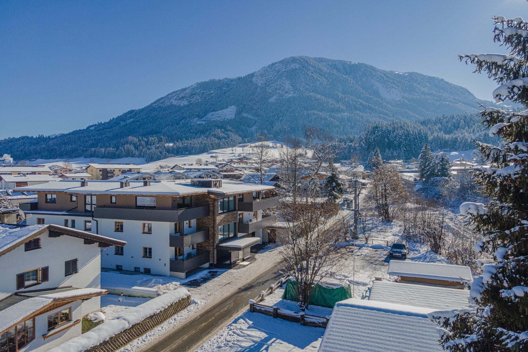 Ein verschneiter Wohnbereich mit modernen Gebäuden, einer klaren Straße und einem Berg im Hintergrund.