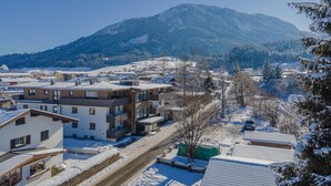 A snow-covered residential area with modern buildings, a clear road, and a mountain in the background.