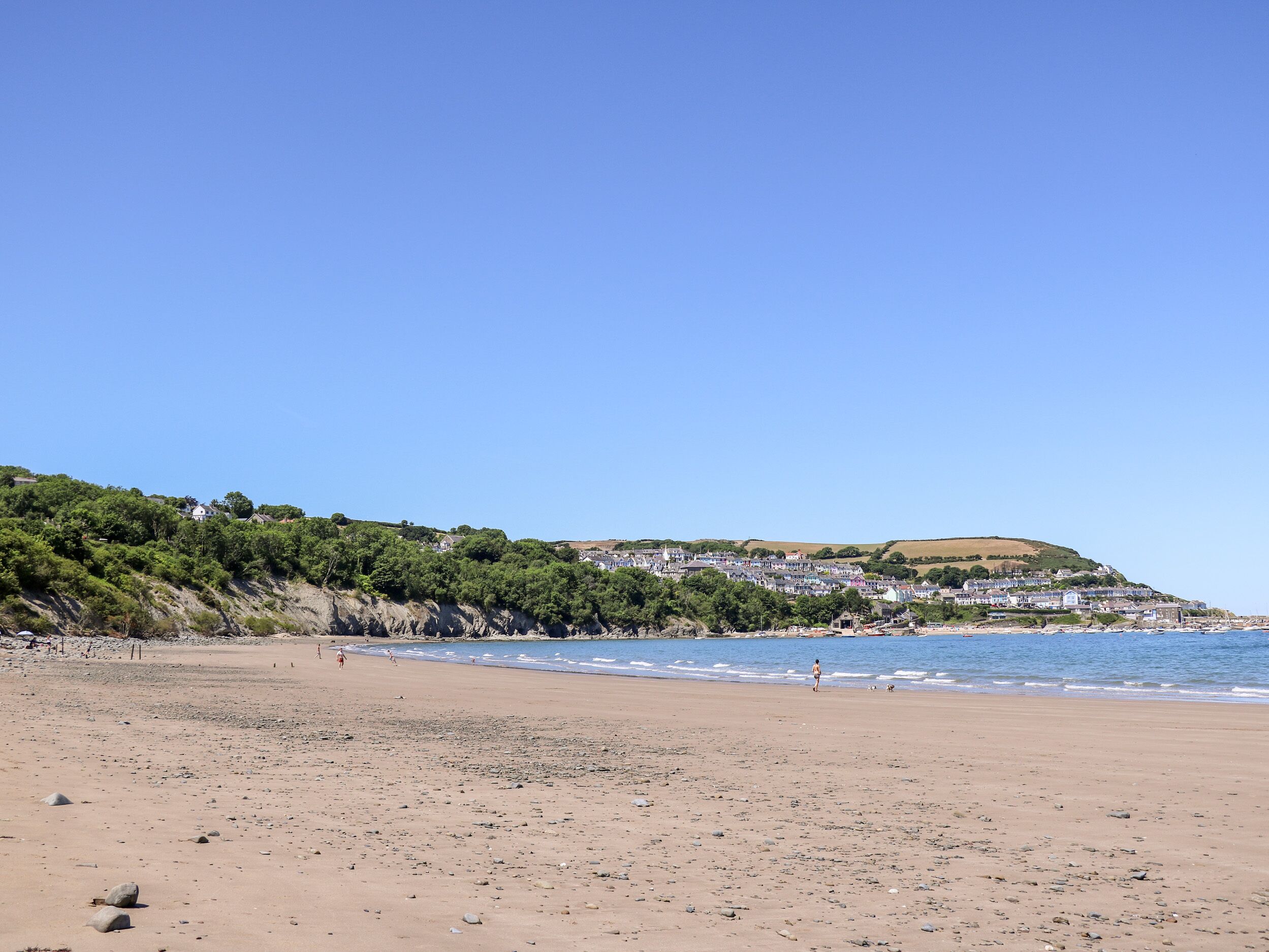 A beach with a clear sky, rocky shoreline, and a town on a hill in the background.