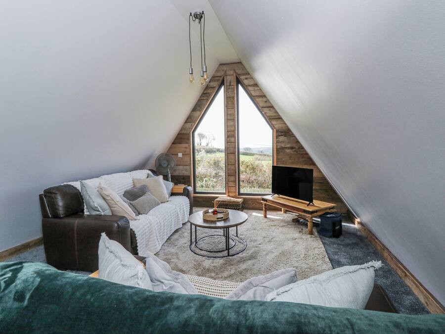 A cozy attic bedroom with a sloped ceiling, a large window, a flat-screen TV, a wooden bench, and a round coffee table.