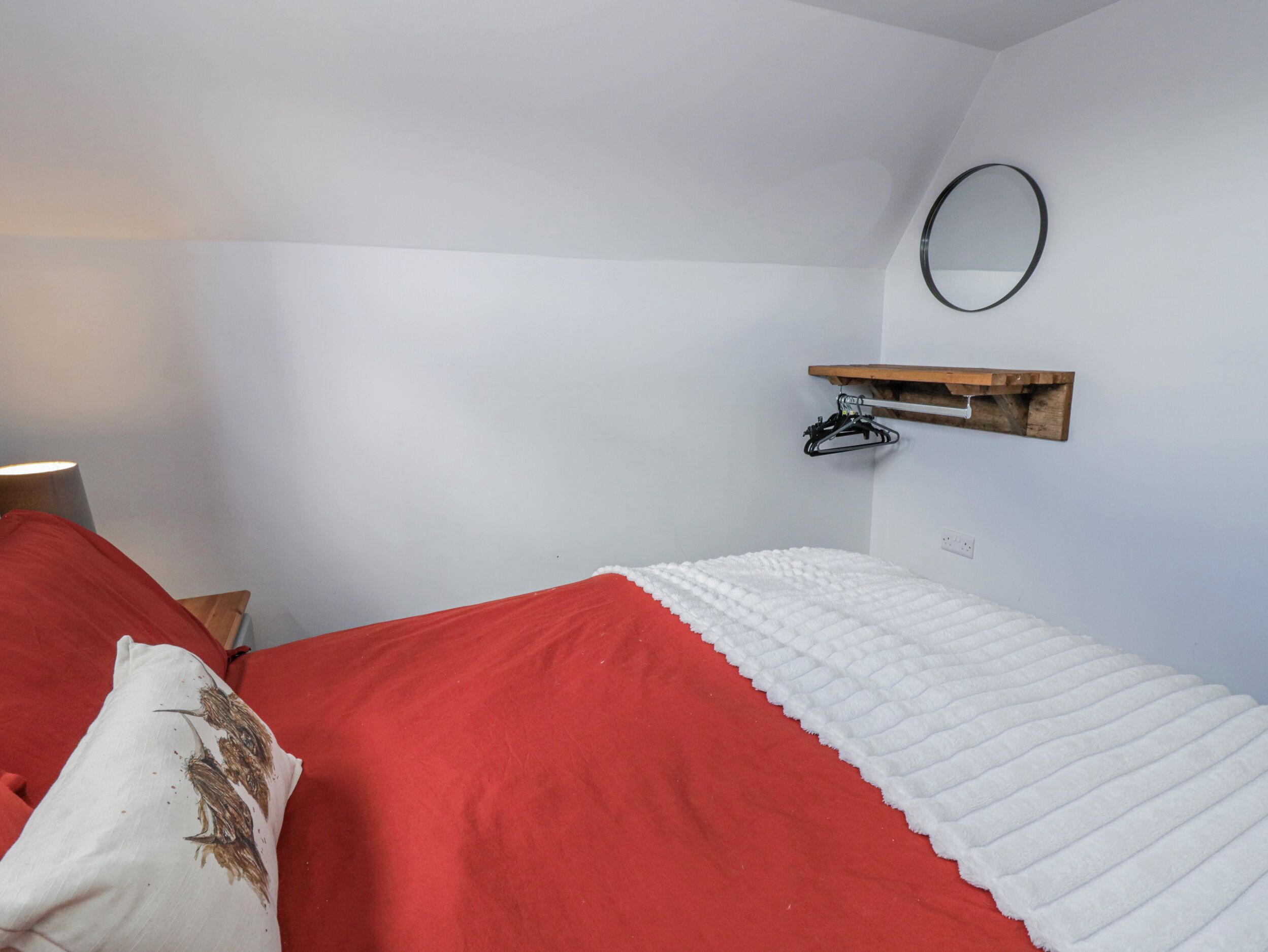 Ein Schlafzimmer mit einem Bett, einem Nachttisch, einer Lampe, einem Wandspiegel und einem Regal mit Kleiderhaken.