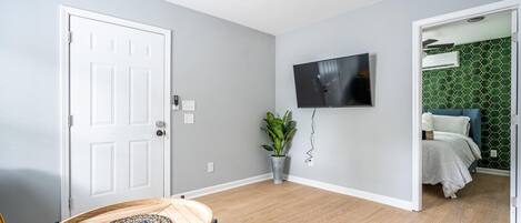 A modern room with a wooden floor, a round table, a potted plant, a flat-screen TV, and a bed with a green patterned wall.