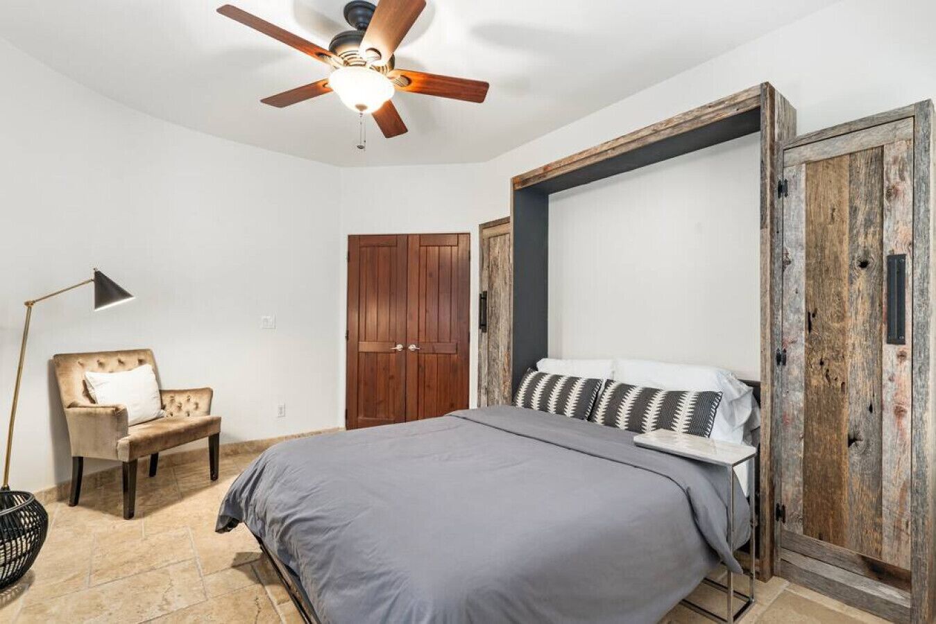 A bedroom with a bed, a chair, a wardrobe, a ceiling fan, and a floor lamp.
