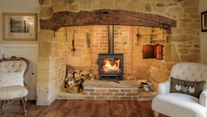 Ein Steinkamin mit einem modernen Holzofen, flankiert von zwei Polsterstühlen und einer rustikalen Holzbalkendecke.