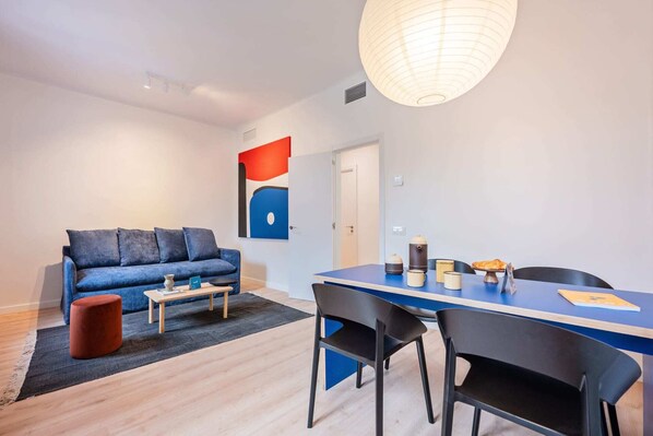 A modern living room with a blue sofa, a coffee table, and a dining table with chairs. There is a white ceiling light and a door in the background.