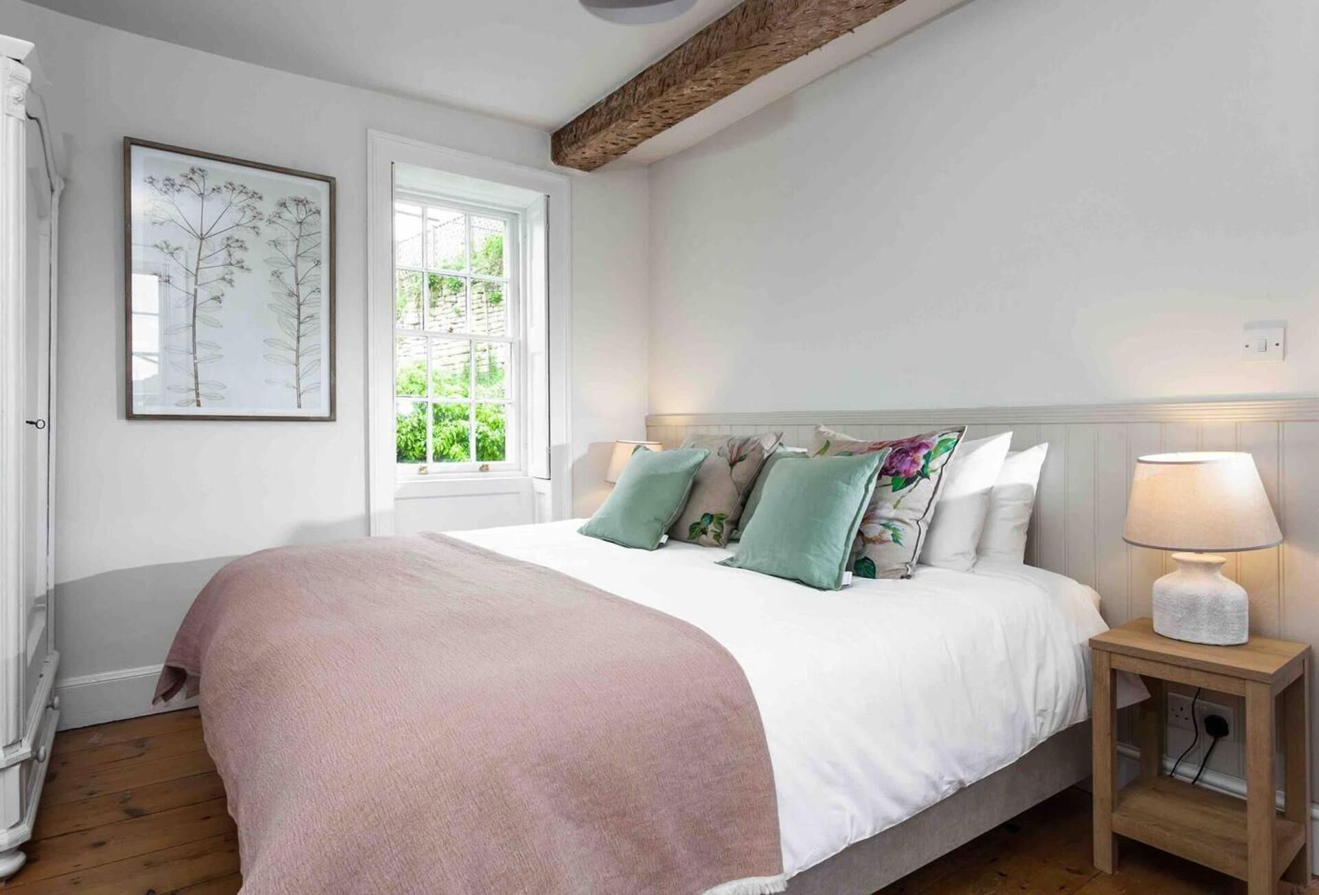 A bedroom with a bed, pillows, a nightstand, a lamp, a window, and a framed botanical print on the wall.