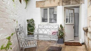A small, white-walled courtyard with a metal bench, a potted plant, and a door leading to a kitchen. - Beautiful Cotswold Cottage in Rodborough, Stroud (Stroud)