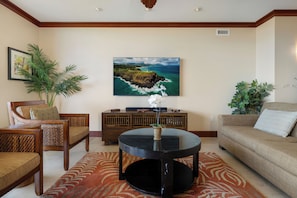 A living room with a round black coffee table, a sofa, two armchairs, a wooden cabinet, and a large framed picture of a coastal landscape.