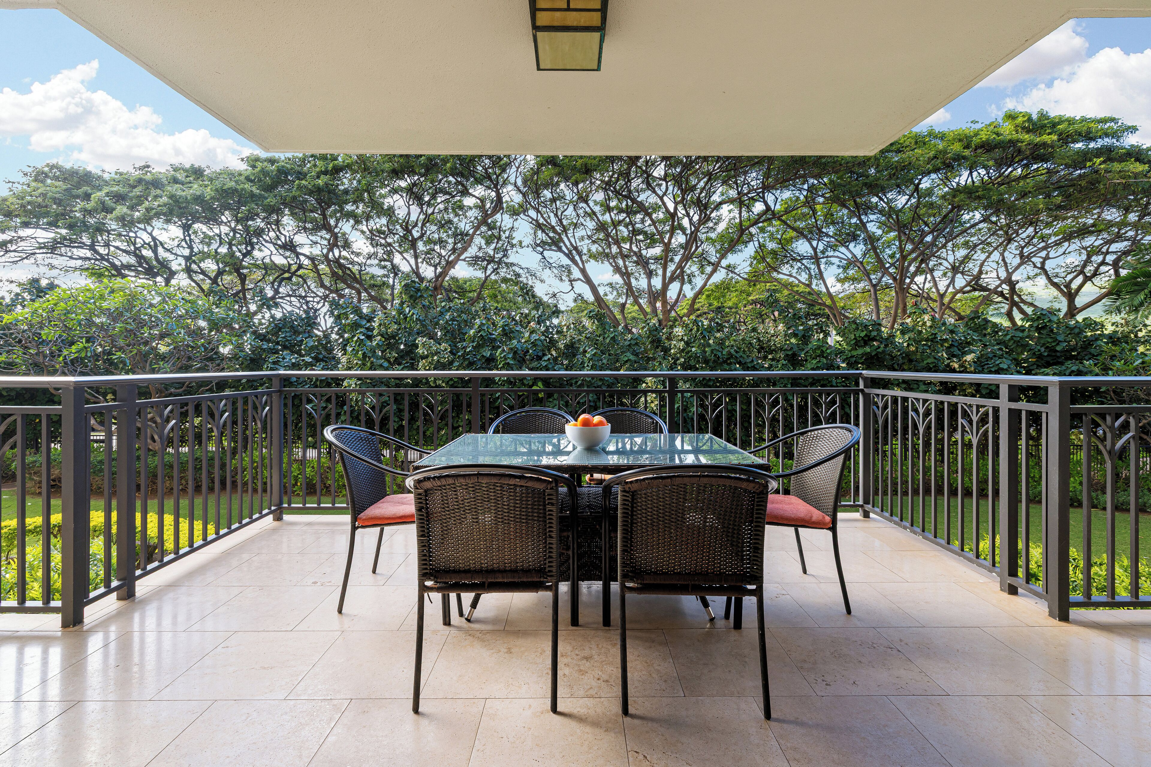 A balcony with a table and chairs, surrounded by a metal railing, overlooking a garden with trees and a clear sky.