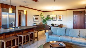 Una cocina moderna con una isla central, taburetes de bar, una zona de comedor con mesa y sillas, y una sala de estar con sofá y mesa de centro.