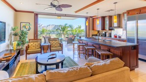A modern living room with a sofa, coffee table, and chairs, a kitchen with a large island and appliances, and a balcony with a view of palm trees and buildings. - Ko Olina Beach Villas B505: Luxe Home w/ Pool & Ocean Views (Kapolei)
