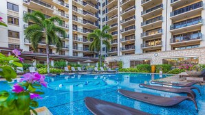 Zona de piscina de un hotel con múltiples sillas de descanso, una piscina central grande y un edificio de varios pisos con balcones.