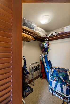 A compact storage closet with a vacuum cleaner, a luggage cart, and a hanging rack with folded clothes.