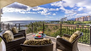 A balcony with wicker furniture, a table with a drink and snacks, and a view of the ocean and buildings.