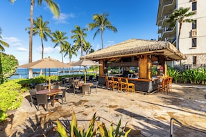 An outdoor bar area with wooden furniture and a thatched roof, surrounded by palm trees and a view of the ocean. - Ko Olina Beach Villas 706: 2BR Villa w/ Panoramic Ocean View (Kapolei)