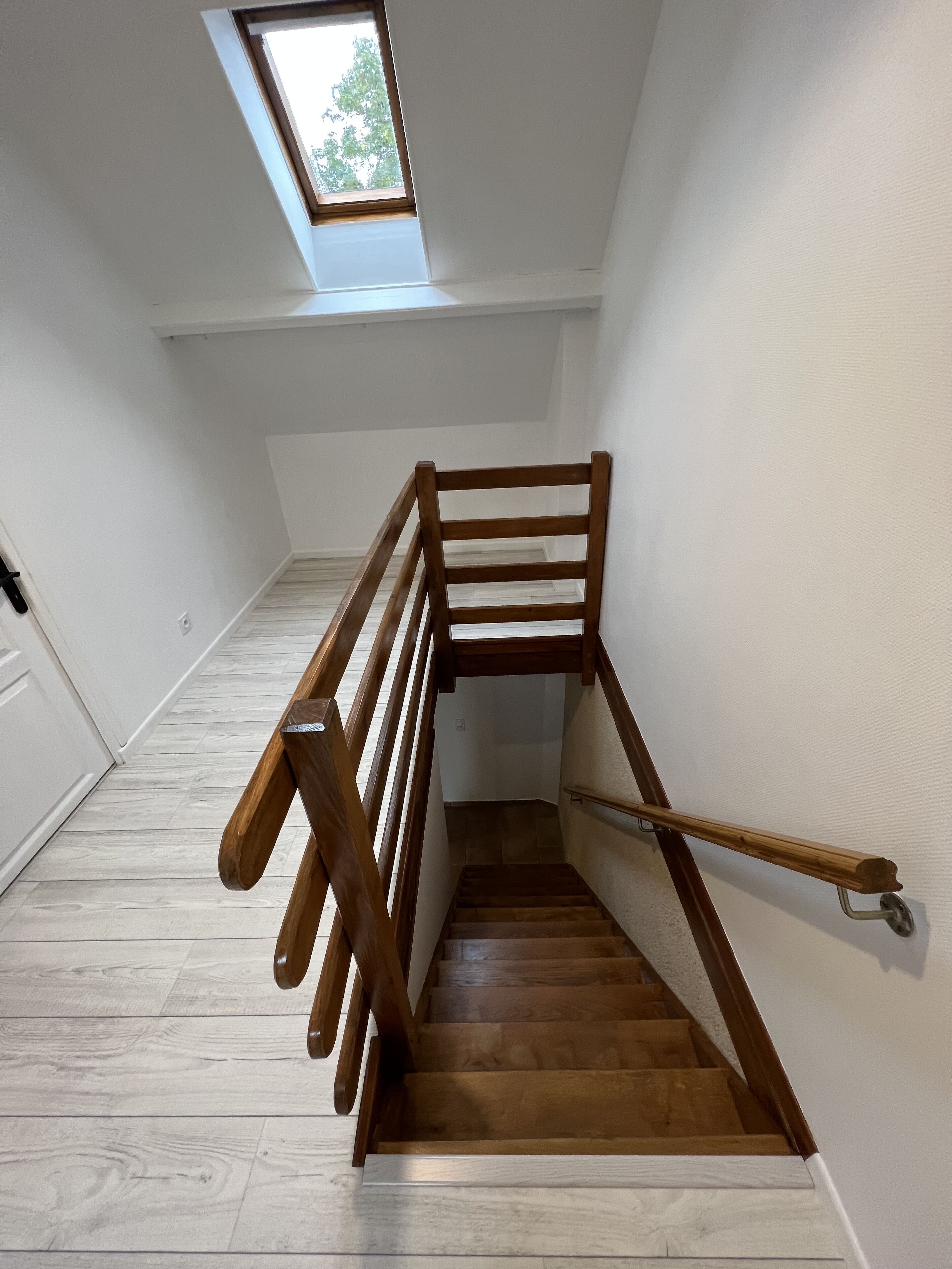 Un escalier en bois avec une rampe, des murs blancs et une lucarne.