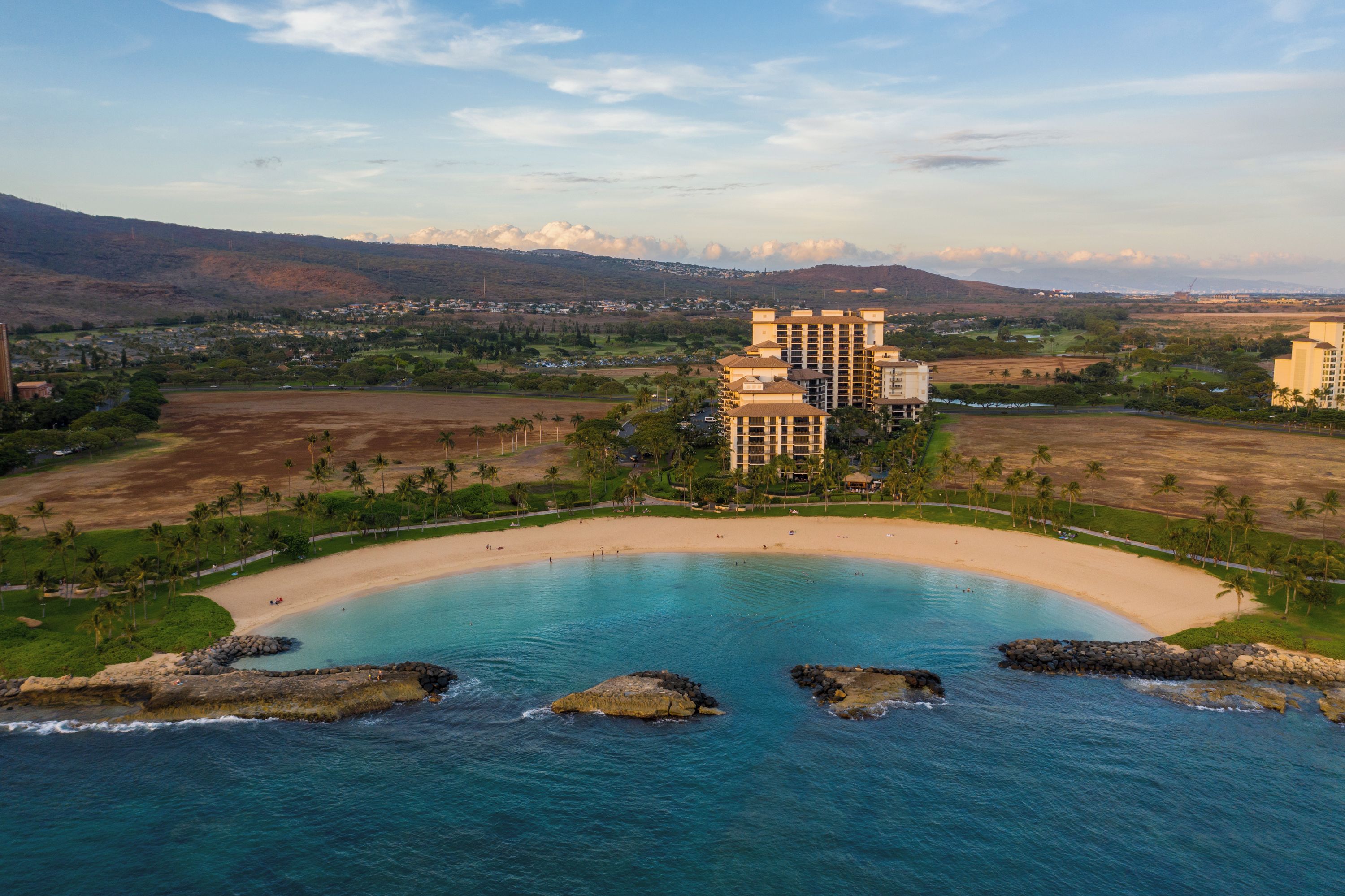 Un resort costero con una amplia playa, un edificio central de varios pisos y vegetación circundante.