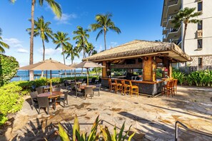 An outdoor bar area with wooden furniture and a thatched roof, surrounded by palm trees and a view of the ocean.
