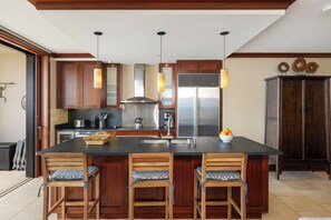 A modern kitchen with a large island, wooden chairs, stainless steel appliances, and pendant lighting.