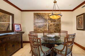 A dining area with a glass table surrounded by wicker chairs, a wooden cabinet, and a large wall art piece. - Ko Olina Beach Villas O805: Chef's Kitchen & Stunning Views (Kapolei)