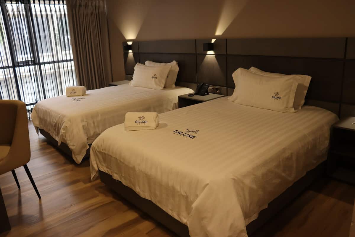 A hotel room with two beds, each with a Giluxe branded pillow and blanket. There is a chair and a nightstand with a phone.