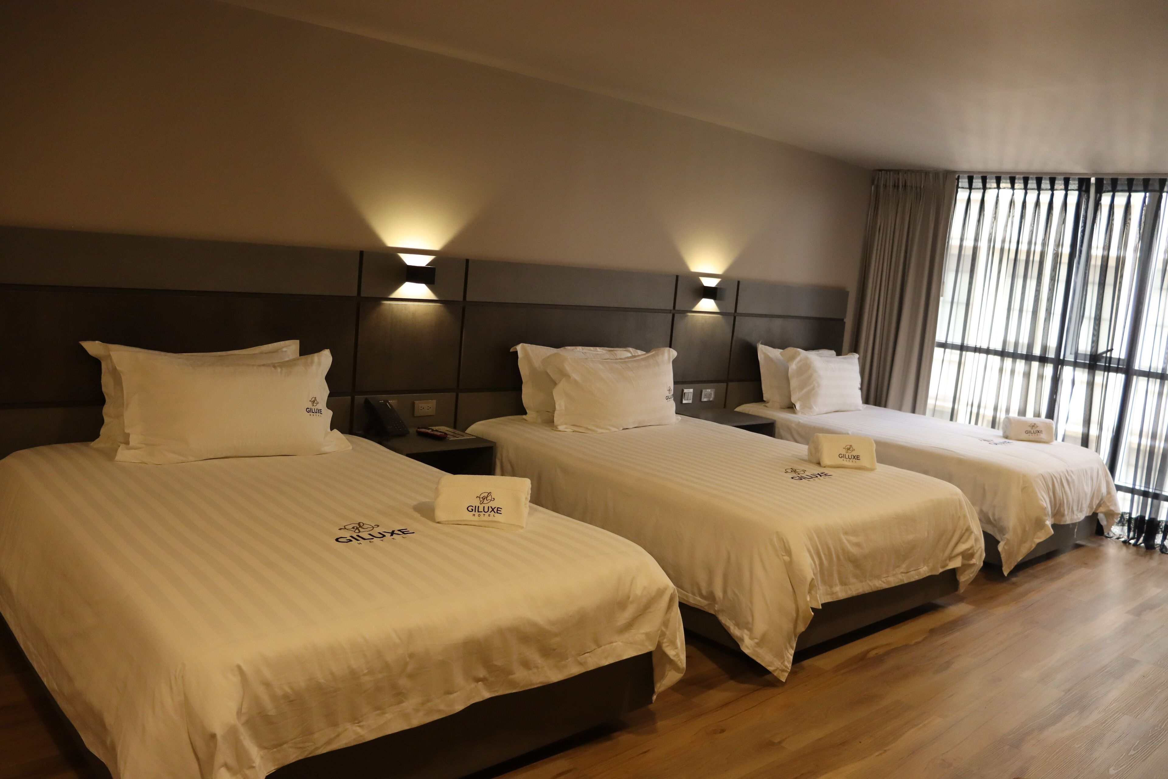 A hotel room with three beds, each with a white bedspread and pillows, and a headboard with a light fixture.