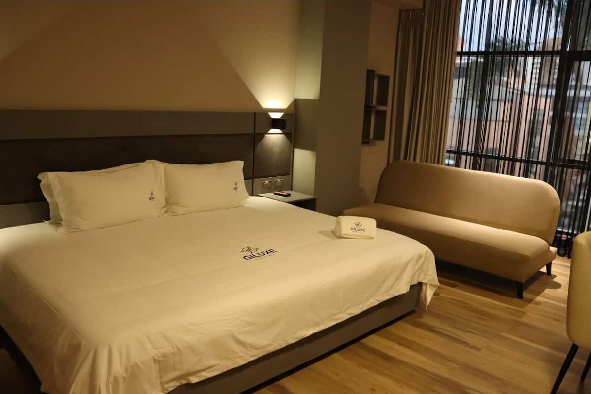 A hotel room with a large bed, a bedside table with a lamp, a sofa, and a window with curtains.