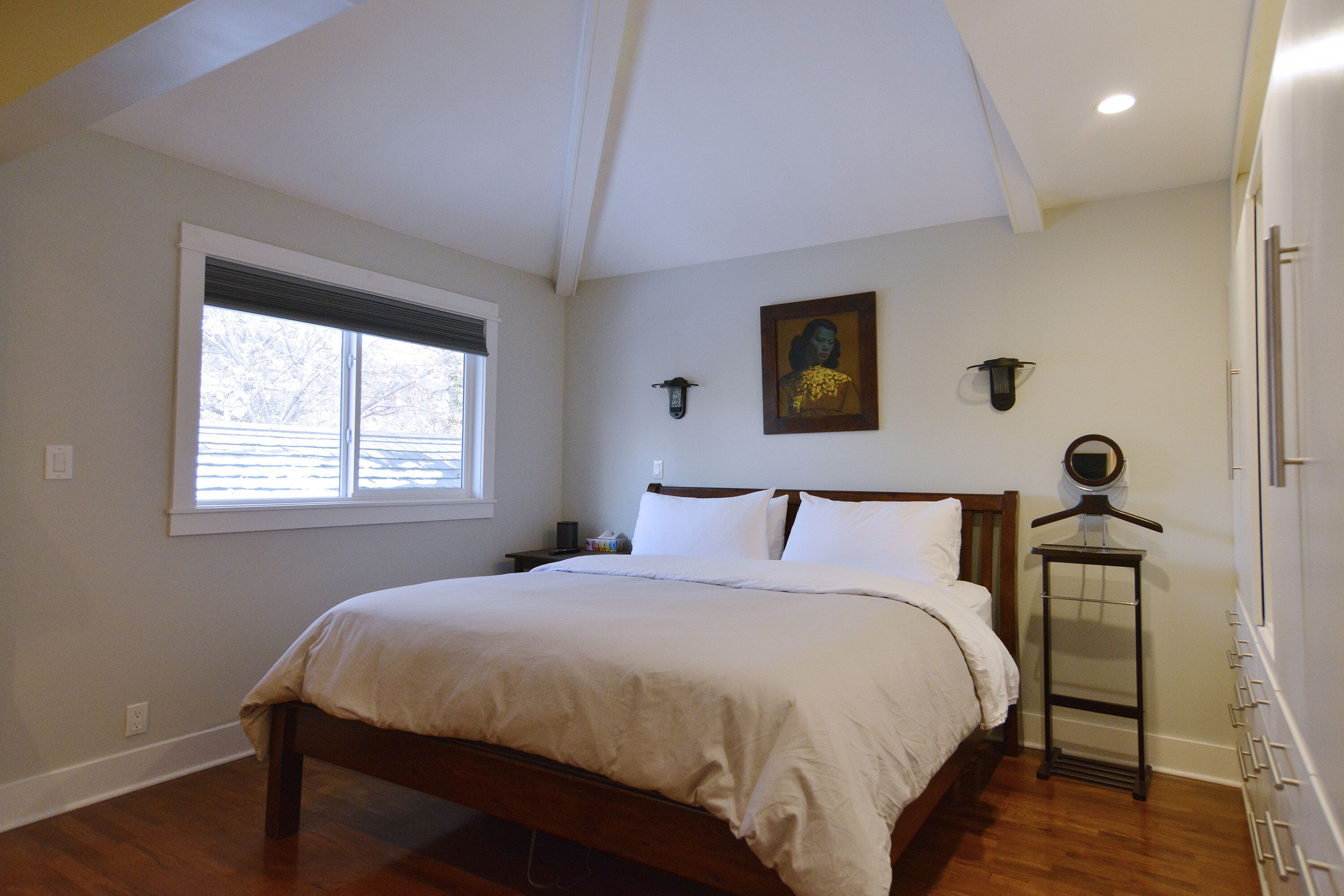 Un dormitorio con una cama grande, una ventana con persianas, un reloj montado en la pared y un cuadro enmarcado.