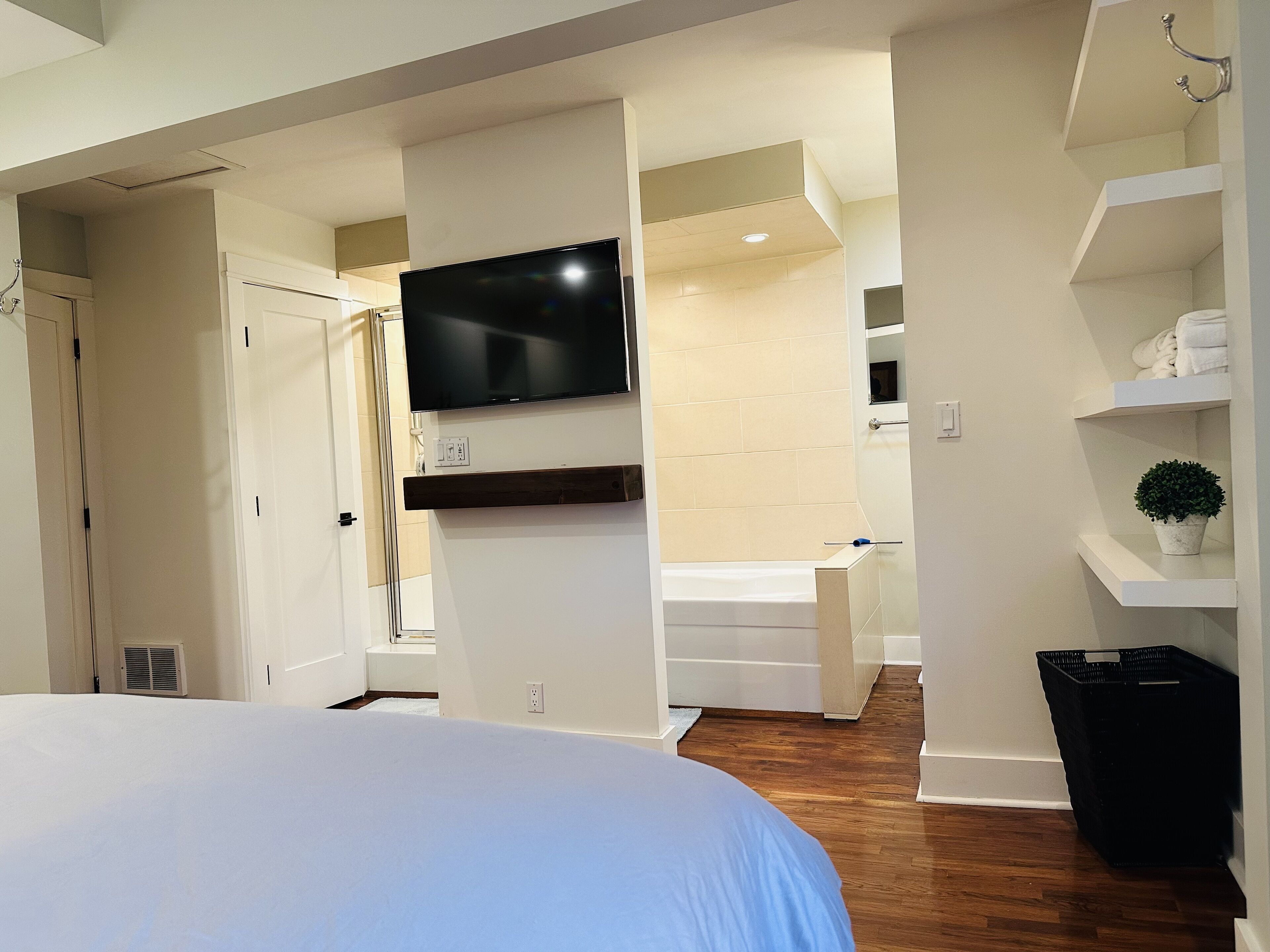 Habitación de hotel moderna con una cama grande, un televisor de pantalla plana en la pared, una bañera integrada y una pequeña planta en maceta sobre una repisa.