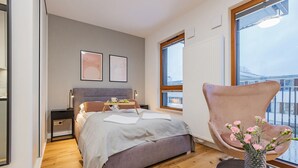 Une chambre moderne avec un grand lit, des tables de chevet, un fauteuil et une vue sur la ville grâce à de grandes fenêtres.