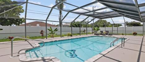 A covered outdoor swimming pool with a metal frame and a clear roof, surrounded by a concrete deck and a white fence.