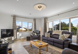 Ein modernes Wohnzimmer mit einem Flachbildfernseher, einer Couch, einem Couchtisch und einem großen Fenster mit Blick ins Freie.