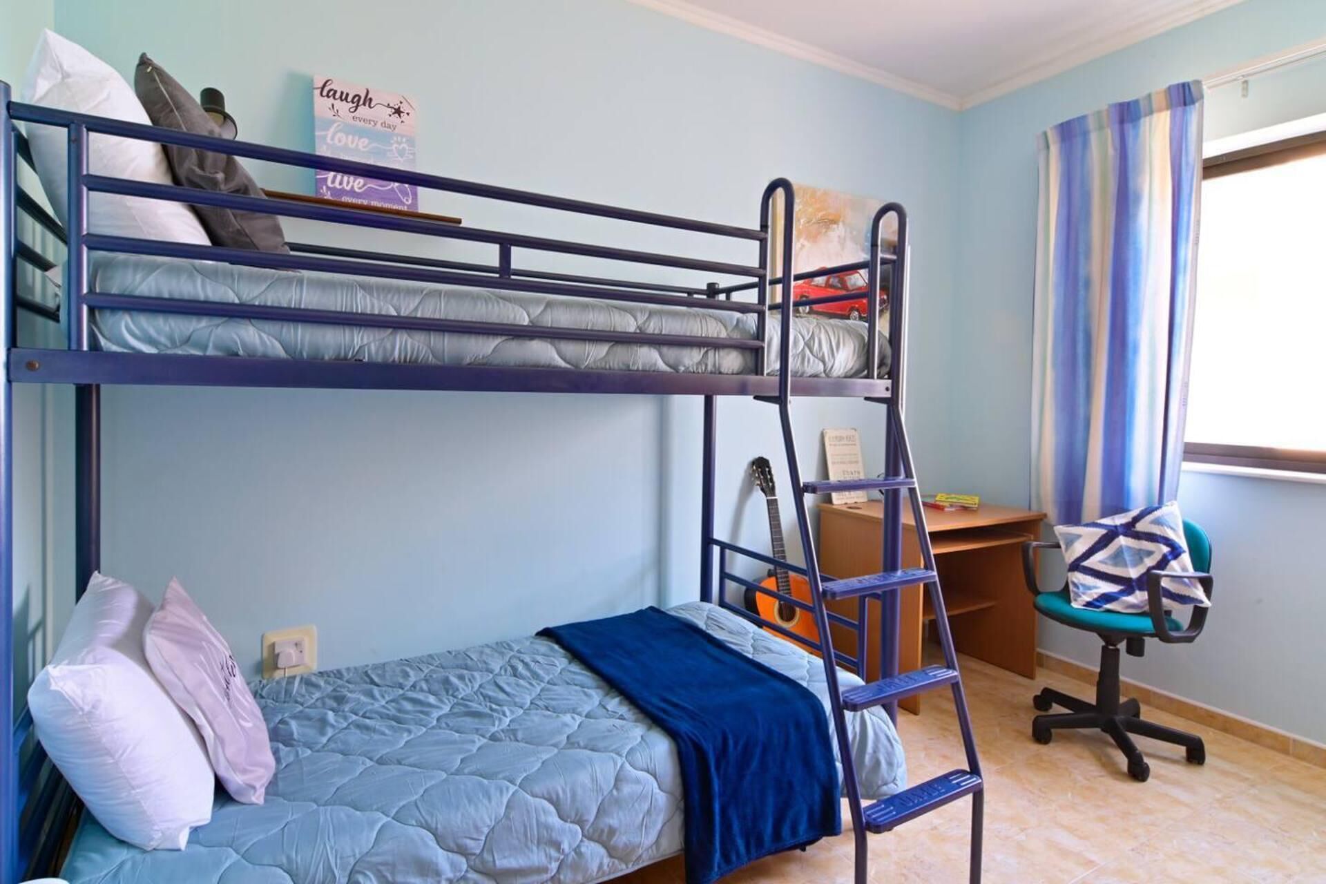 A bunk bed room with a desk and chair, a guitar, and a window with curtains.