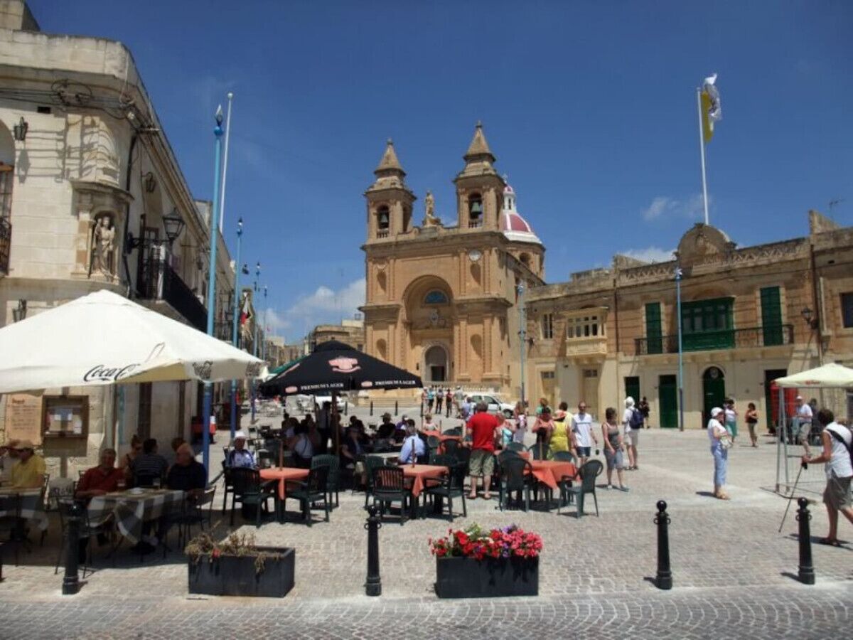 Un'area caffè all'aperto con tavoli e sedie, un edificio importante sullo sfondo con un campanile e un cielo azzurro limpido.