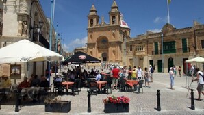 An outdoor café area with tables and chairs, a prominent building with a bell tower in the background, and a clear blue sky.